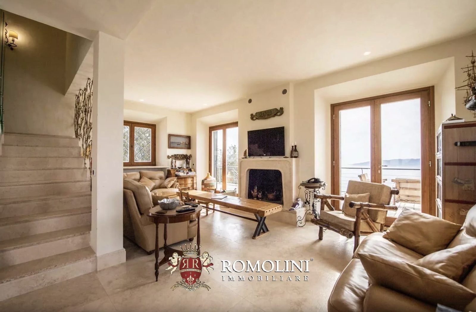 Ground-floor living room with stone fireplace, leather sofas, and French doors opening to a terrace with sea views