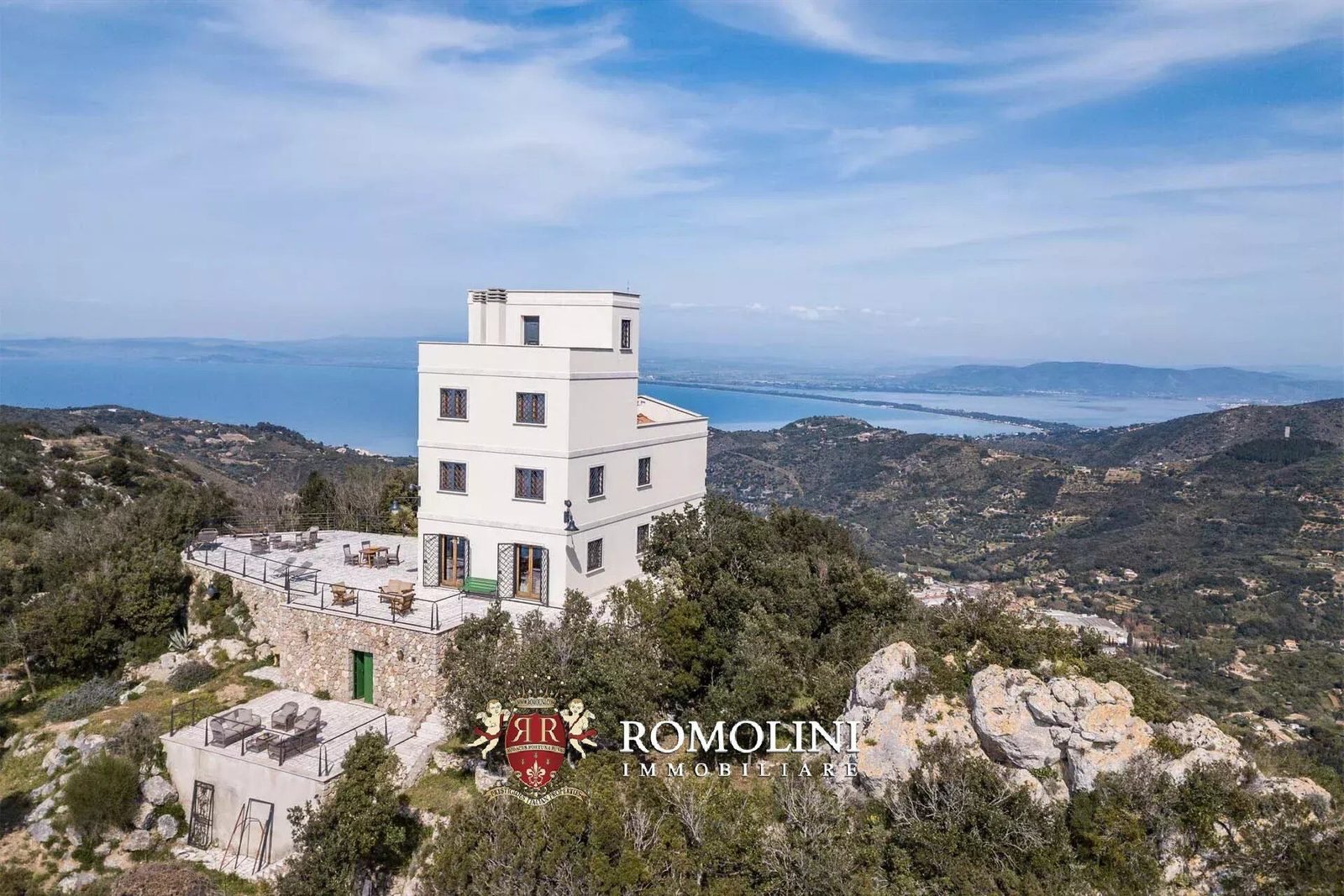 Four-storey white tower villa above stone retaining walls with the Orbetello lagoon and Tuscan coastline in the distance
