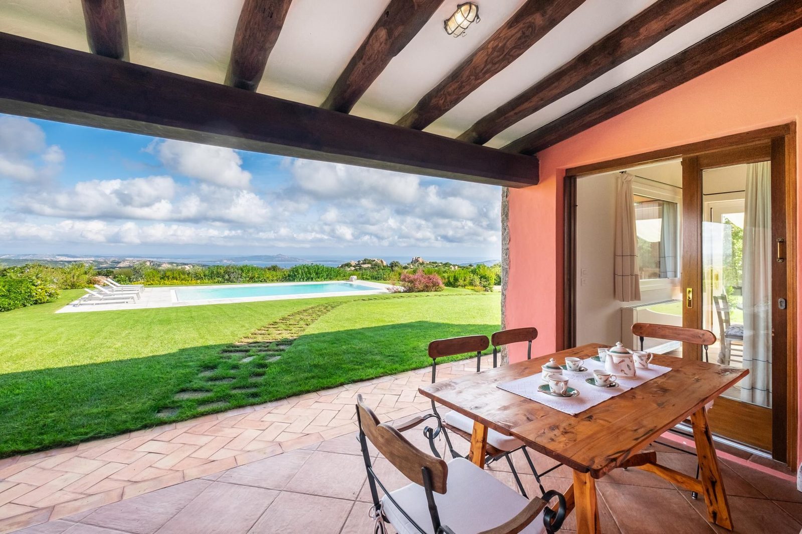Covered terrace with dining table overlooking the pool, lawn, and coastline of a three-bedroom villa in Cannigione, Costa Smeralda, Sardinia